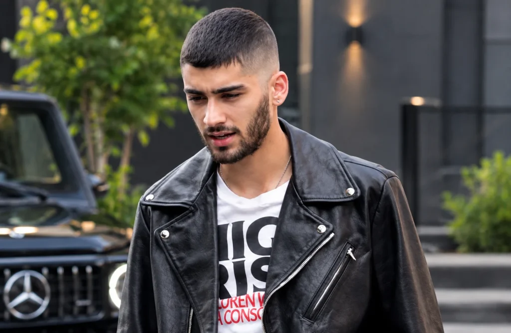 Young man wearing a black leather jacket and white graphic T-shirt, featuring a short buzz cut with a clean fade and neatly trimmed beard, standing outdoors in an urban setting.